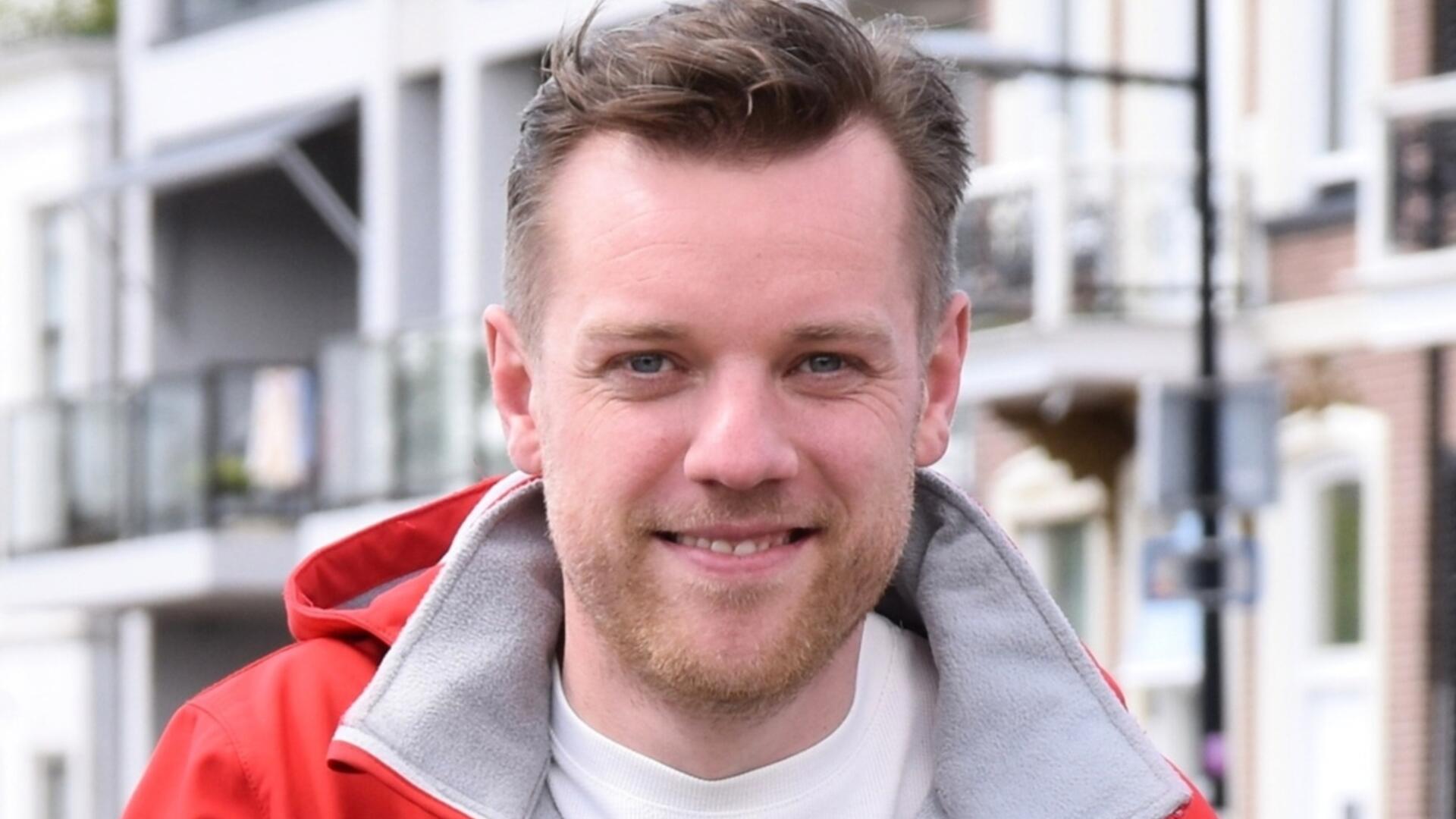 Foto van Willard Bouwmeester tijdens een campagne actie. Willard draagt een rode PvdA jas, op de achtergrond zie je appartementen gebouwen. De foto is genomen in de buurt van het centrum van Assen.